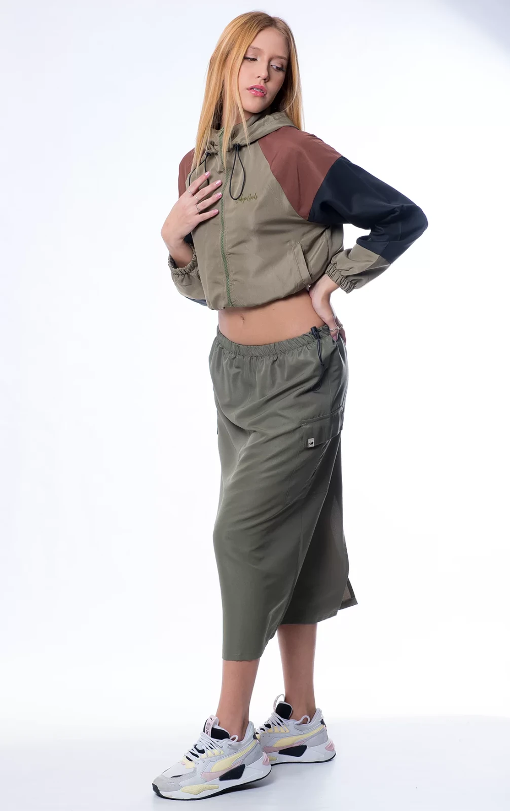 Menina com um look com saia midi, jaqueta corta vento verde militar e tênis no estilo esportivo